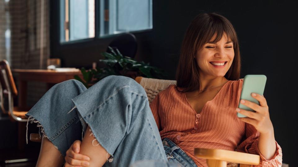 Portrait of a beautiful cheerful young Caucasian businesswoman wearing a pink blouse and jeans using a mobile phone while sitting casually in a chair at home.