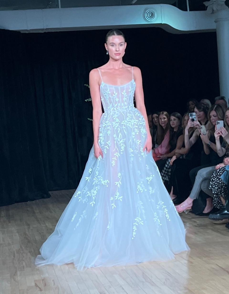 A bride walks down a runway in a sheer, floral dress.