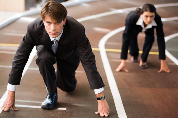 A man and a woman, dressed in dark business attire, in starting positions on a running track.