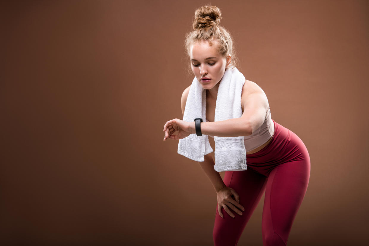 Tired young blond sportswoman in activewear looking at fitbit on her wrist while doing difficult physical exercise against brown background