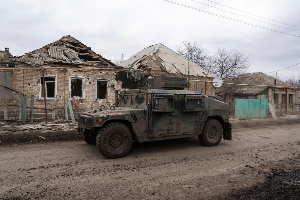 A muddy armoured vehicle in front of destroyed one-story homes against a grey sky