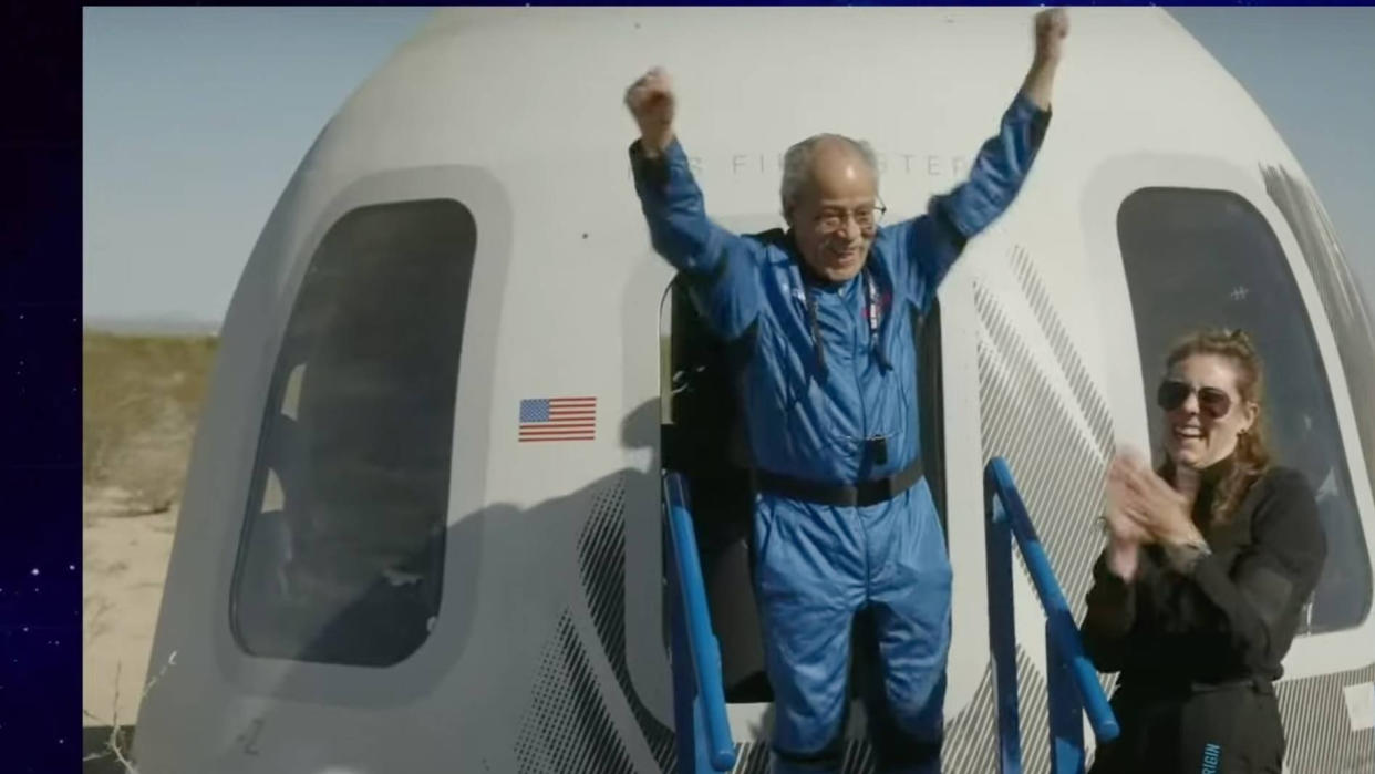  An older man in a blue flight suit emerges from a white space capsule with arms raised in triumph. 