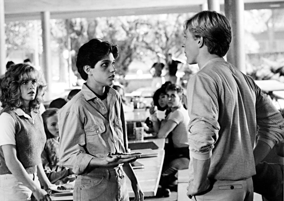 Three actors in a classic film scene, one holding a book and engaging in a conversation