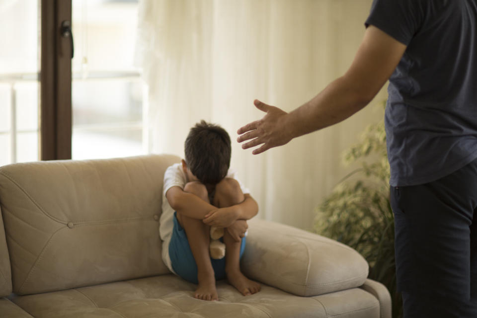 A young boy, upset and curled up on a couch, covers his face with his arms while an adult stands nearby with a hand extended