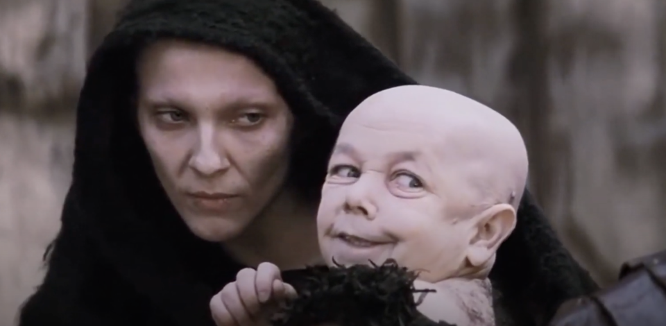 A cloaked figure holds a toddler who is wearing a sinister grin