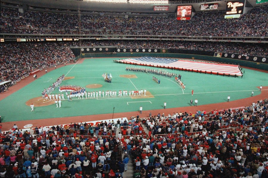 Team members of the Philadelphia Phillies and the Atlanta Braves line the base paths as a giant flag stretches across the outfield before the National Anthem at Veterans Stadium in Philadelphia, Oct. 6, 1993. (AP Photo/Amy Sancetta)