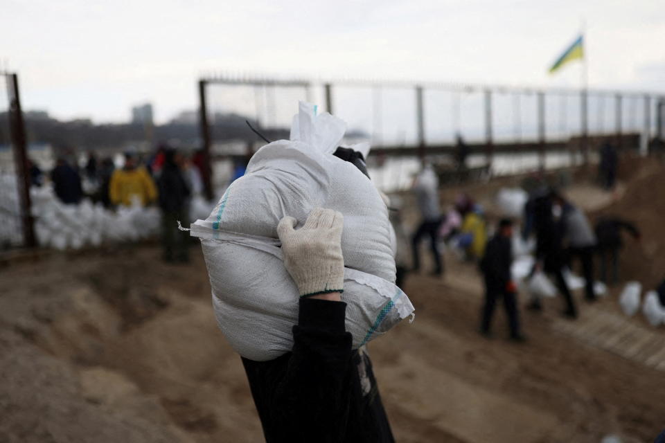 A man carries sandbags on his shoulder near a beach. 