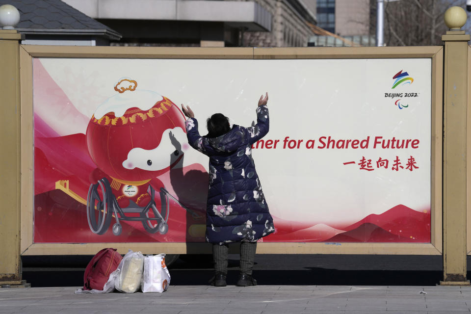 A traveler stretches out as she enjoys a sunny day near an advertisement for the Beijing 2022 Winter Olympics at the Beijing railway station in Beijing, China, Friday, Jan. 28, 2022. The Beijing Winter Olympics is coinciding with the Chinese Lunar New Year and Chinese authorities have called on the public to stay where they are instead of traveling to their hometowns for the year's most important family holiday. (AP Photo/Ng Han Guan)