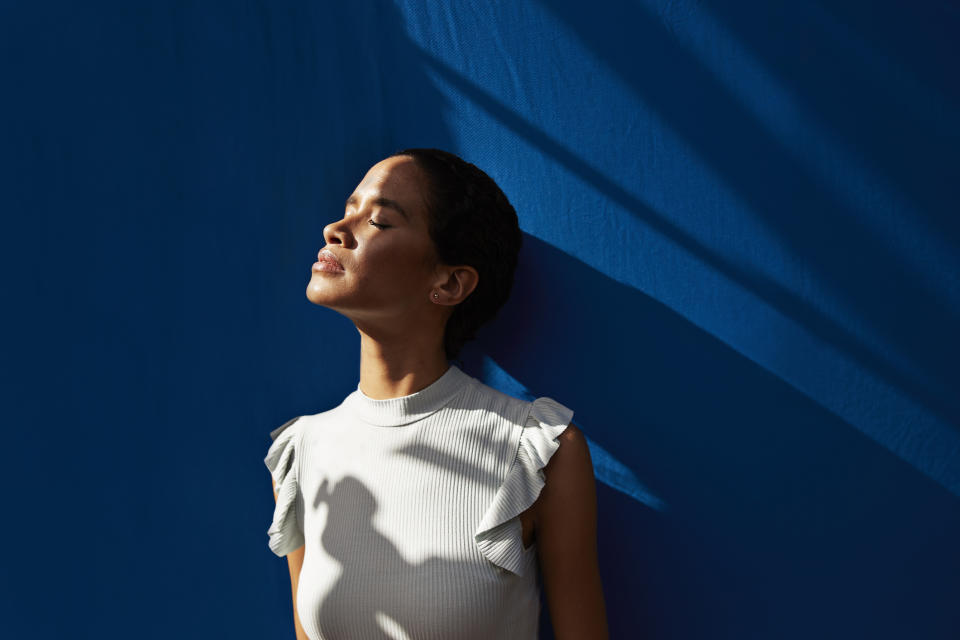 A person stands against a backdrop with eyes closed, basking in sunlight, wearing a sleeveless, ruffled high-neck top