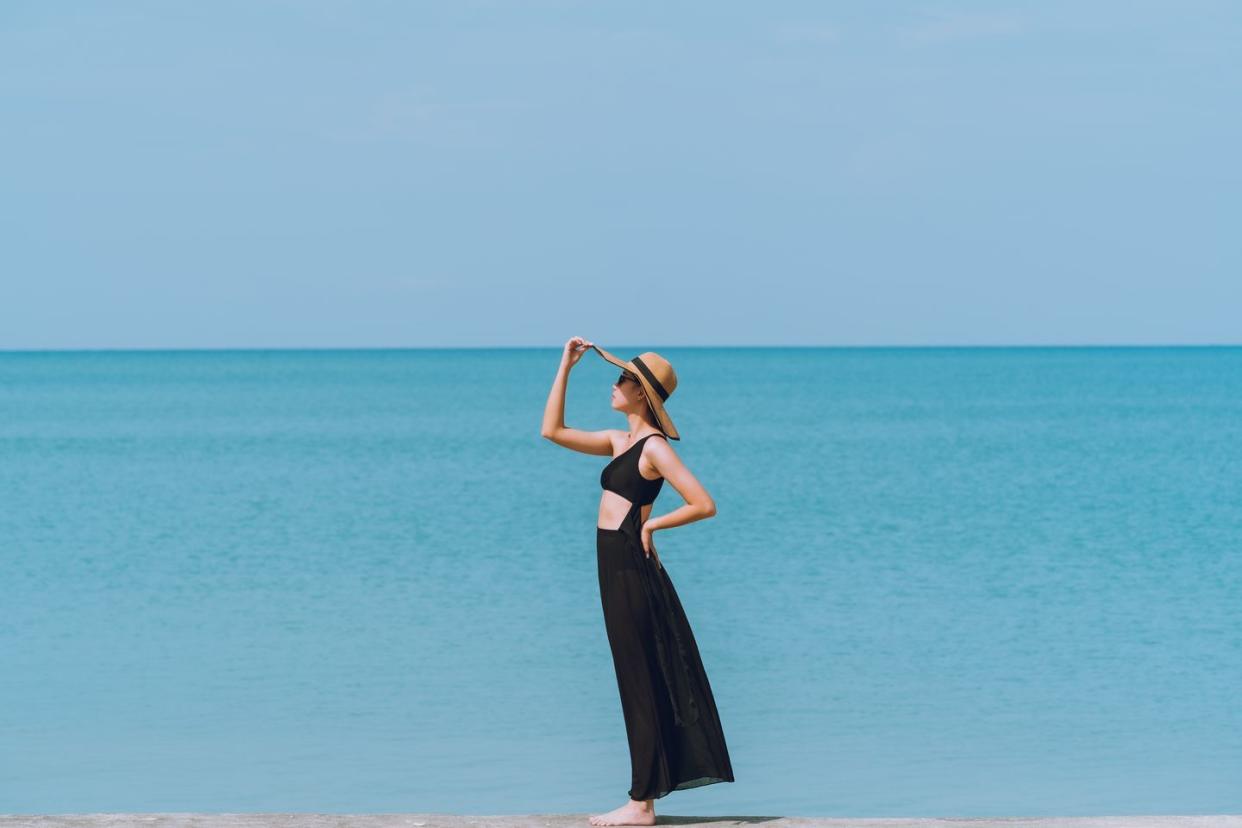 a woman in a black bikini, wearing a long black skirt and wearing a hat, stands by the sea and the sky is blue