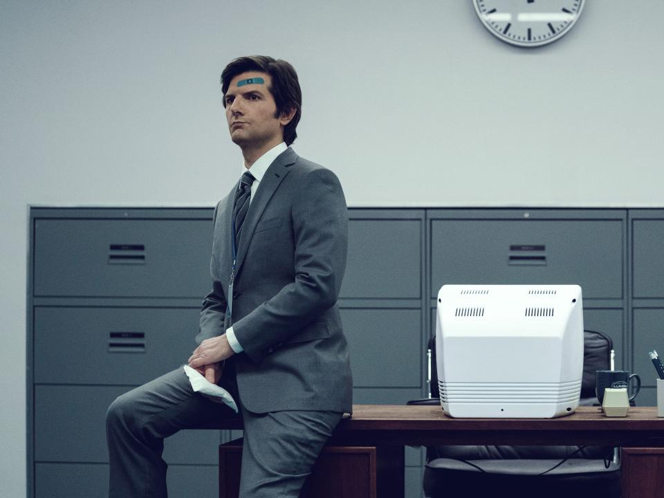 adam scott as mark in severance, sitting on a desk with an older-looking computer and a mid-height row of file cabinets in the background