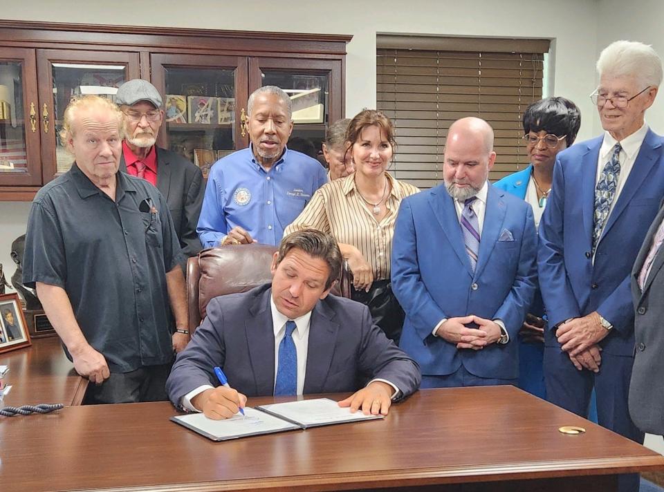 Gov. Ron DeSantis (seated), surrounded by lawmakers, victims and others in the Florida Capitol, signs a bill allowing victims of abuse at two notorious and now closed boys schools in the state to be paid reparations, Friday, June 21, 2024.