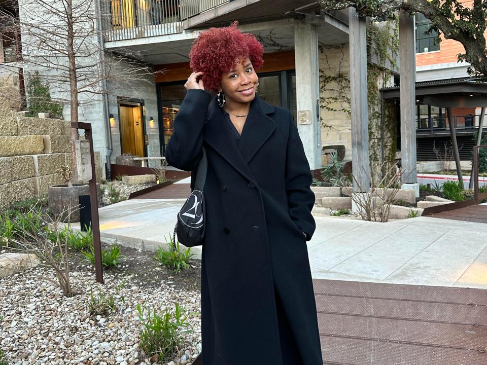 A woman with red curly hair, silvery hoop earrings, and a black coat stands in a courtyard with stones and plants.