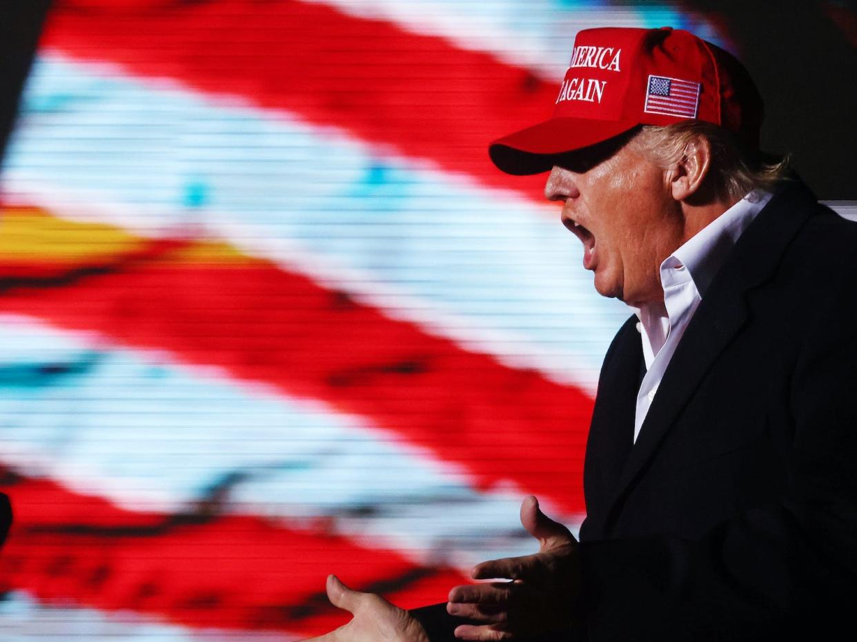 Donald Trump yells in front of an American flag while wearing a MAGA hat.