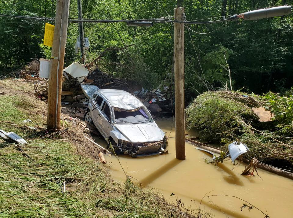 A car head first in muddy water, its roof dented, with what appears to be its door hanging from a power line some feet away.