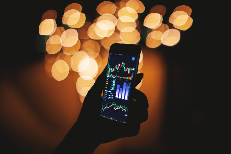 Silhouette of female hand holding smart phone in the dark with stock trading on display with light bokeh blurred background, Mobile business concept