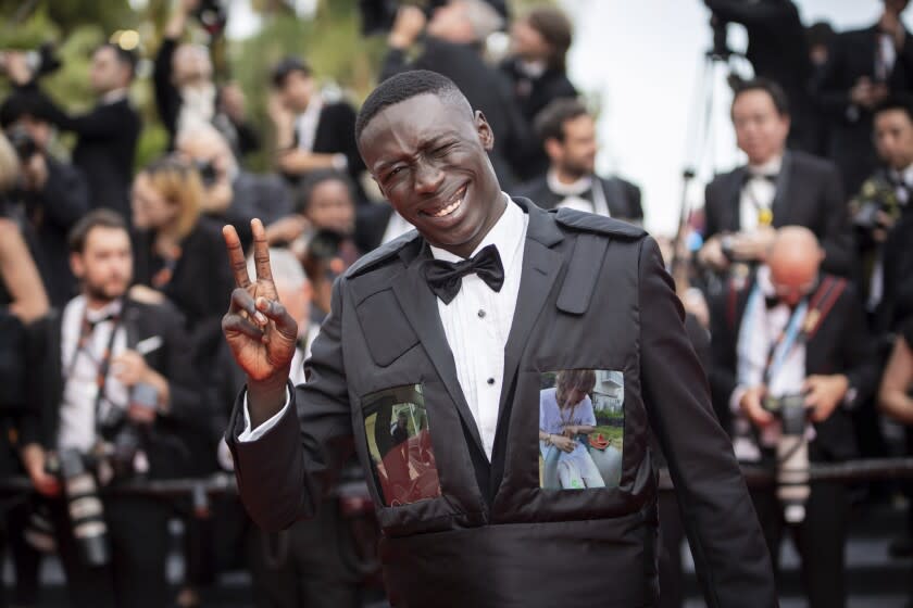 A man in an unusual vest over a tuxedo waves a peace sign to photographers at a movie premiere