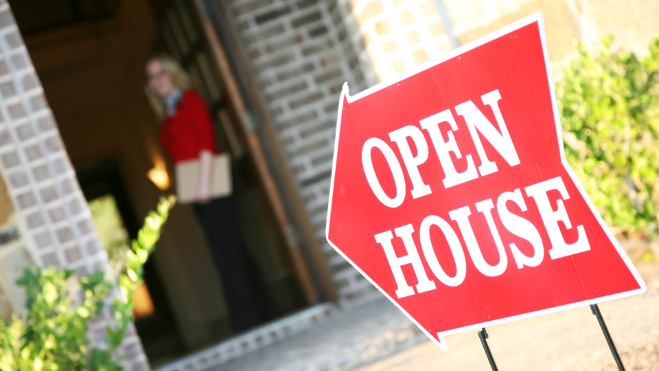 A red 'Open House' sign pointing towards an open door where a real estate agent stands.