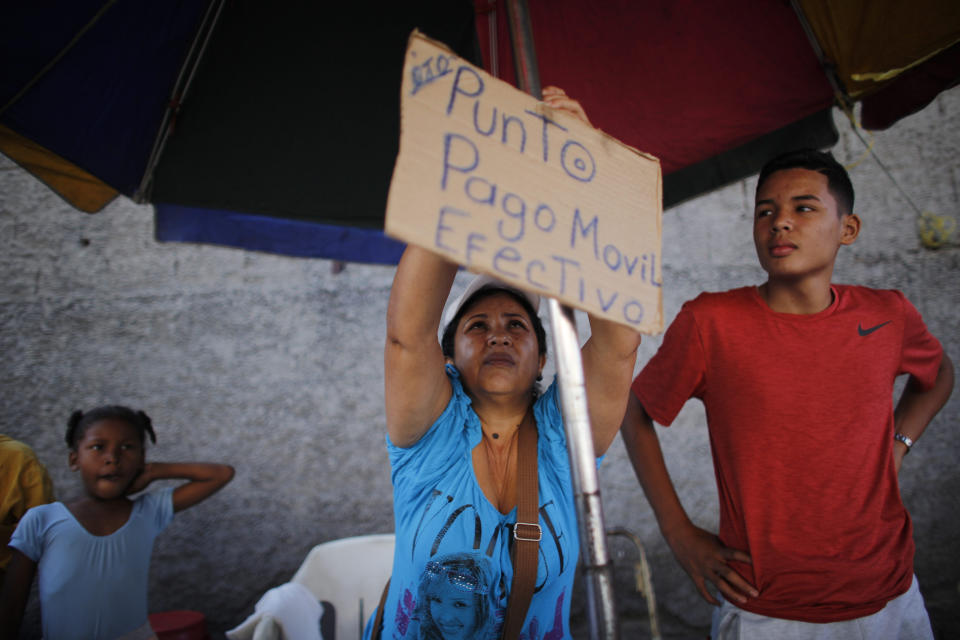 In this Oct. 5, 2019 photo, Daixy Aguero puts up a sign that reads in Spanish "Point of sale, mobile payment and cash," at her stand where she sells beauty products, at a market in Caracas, Venezuela. It's a side-gig that allows her to keep her dream job as a preschool teacher. (AP Photo/Ariana Cubillos)