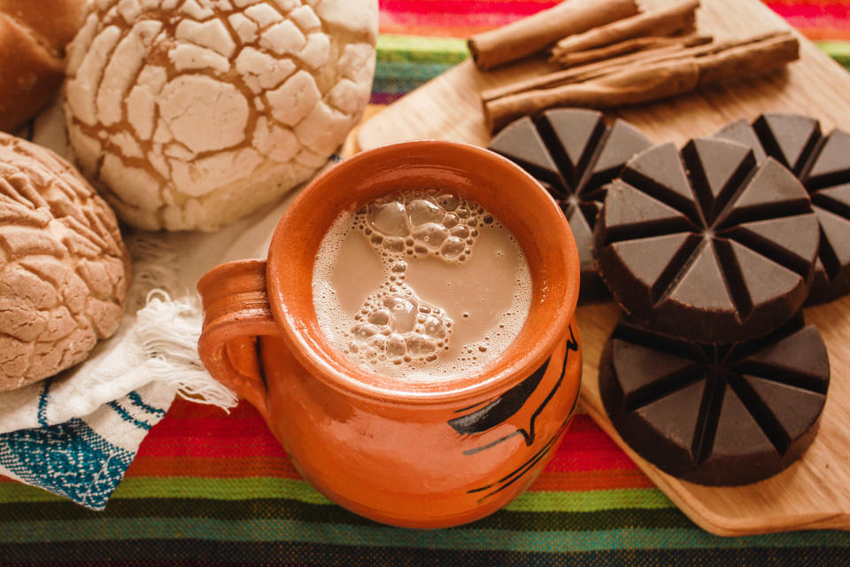 a cup of hot chocolate next to chocolate pieces, cinnamon, and conchas