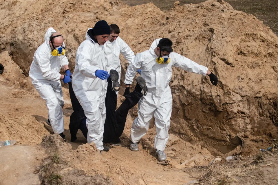 Several men, dressed in white and some wearing masks, drag a body by the legs out of a ditch.