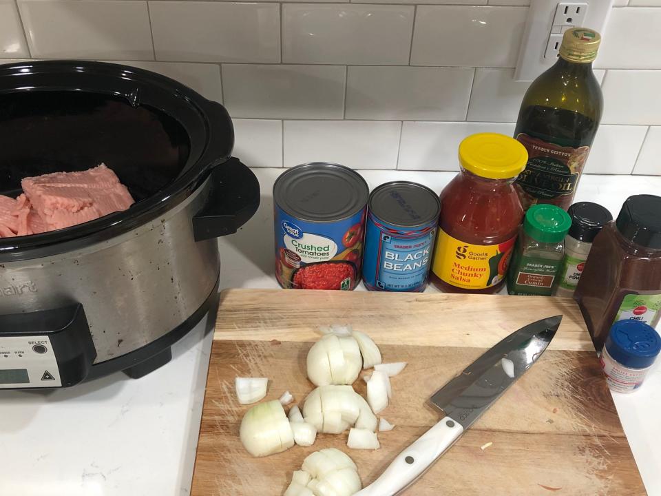 A Crock-Pot filled with uncooked turkey, cans of crushed tomatoes and black beans, a jar of medium chunky salsa, a bottle of olive oil, seasonings, and a cutting board on a white counter. On the cutting board is a knife with sliced onions.