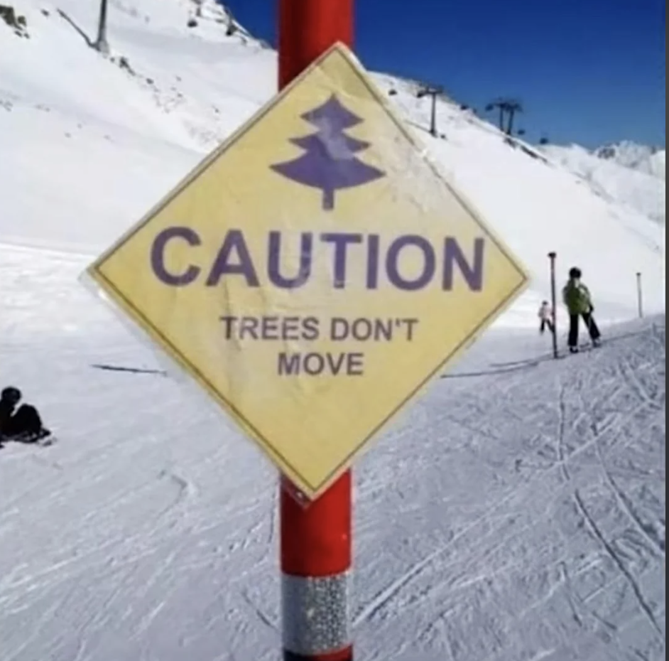 A caution sign on a ski slope reads "TREES DON'T MOVE" with a skier and snowboarder in the background