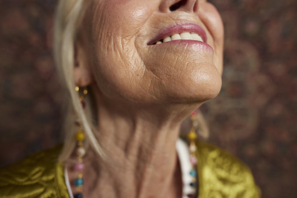 Close-up of an elderly woman smiling, showing her wrinkles. She is wearing colorful beaded earrings and a textured top