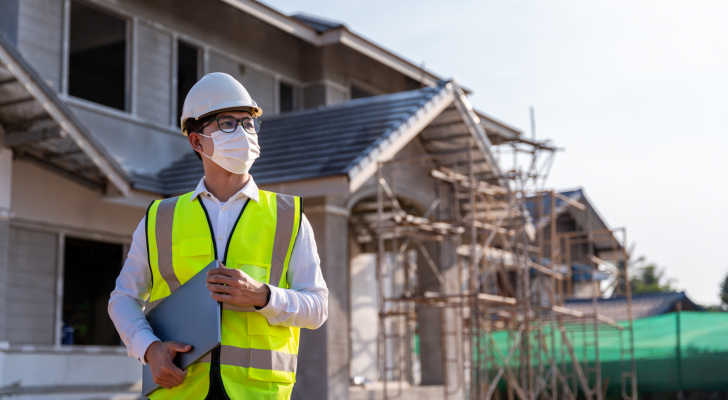 A photo of a man with a clipboard in front of a home under construction.