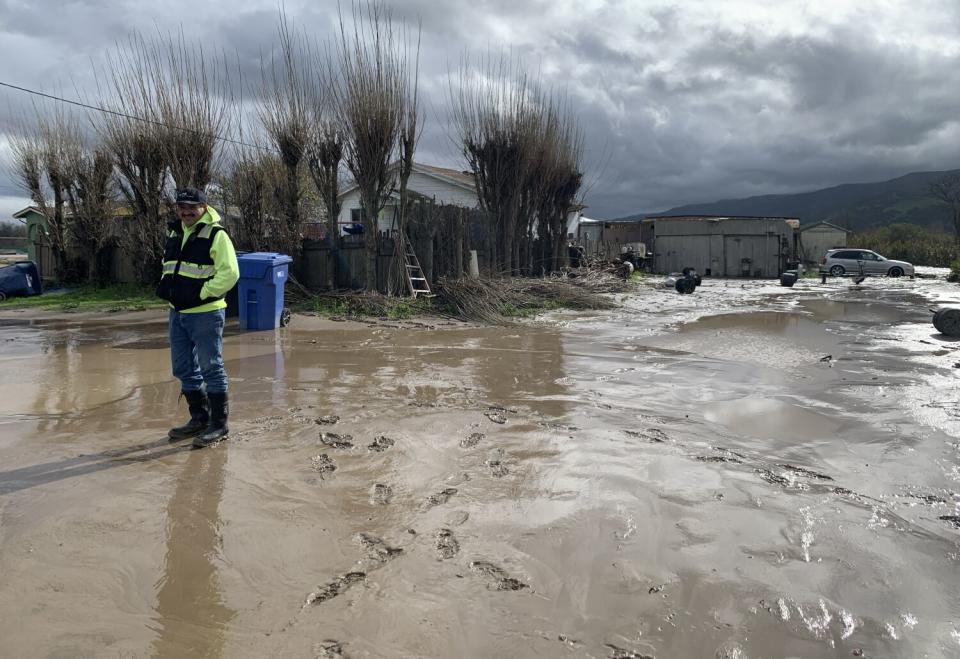 A man in knee-high rubber boots stands amid mud. Behind him are a house with a brown fence and outbuildings.