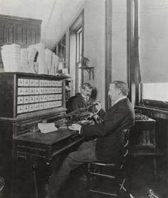 an old black and white photograph showing a man seated at a wooden desk-like machine looking at a bank of indicator dials