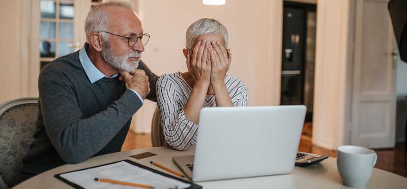 An elderly couple look at a laptop screen, the wife covers her face.