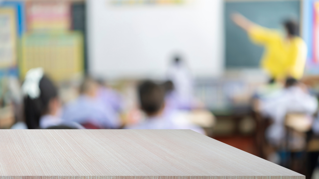 A blurred perspective from a student's desk in the rear of the elementary school classroom showing a teacher point to a whiteboard