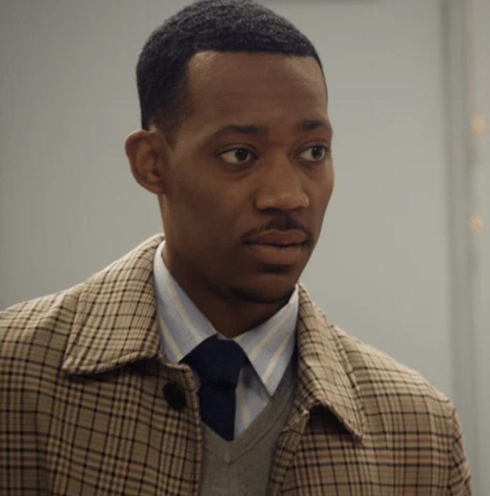 Tyler James Williams dressed in a plaid jacket, blue shirt, and tie stares calmly ahead