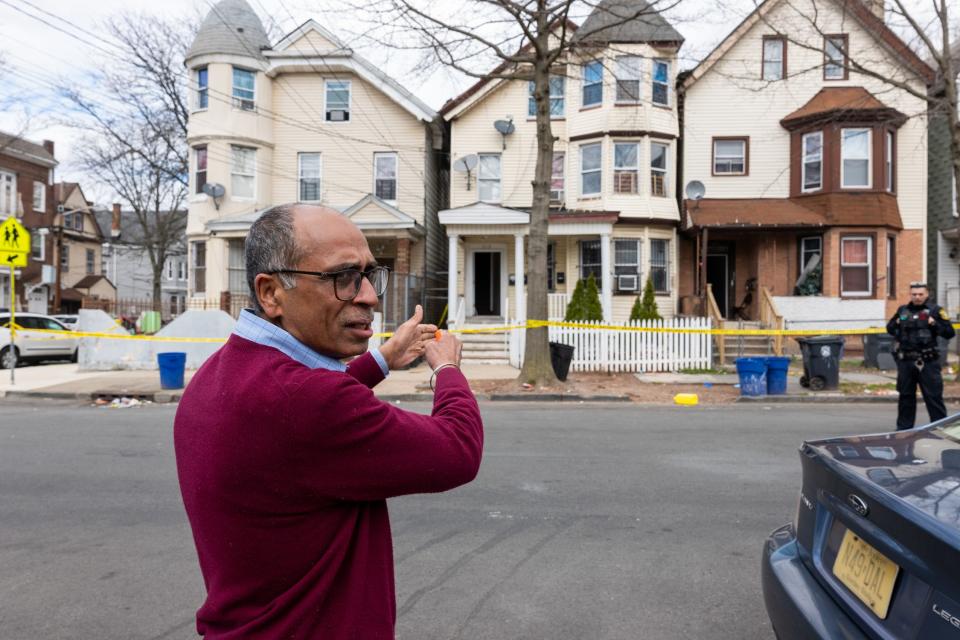 man with concerned expression wearing red sweater gestures at a row of homes blocked off with yellow tape