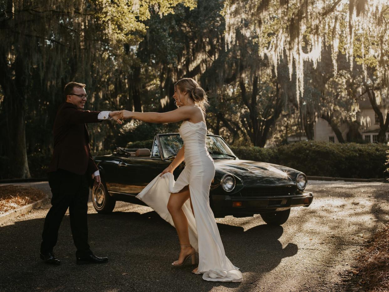 A bride and groom dance together in their wedding attire in front of a convertible.