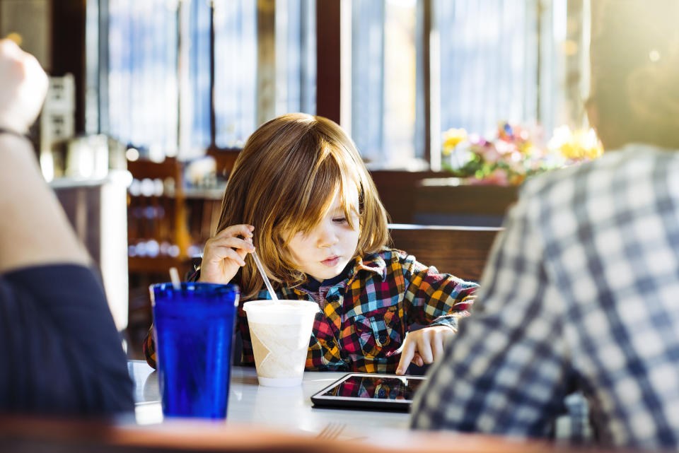 A child with long hair wearing a plaid shirt uses a tablet while sitting at a table with two adults in a restaurant