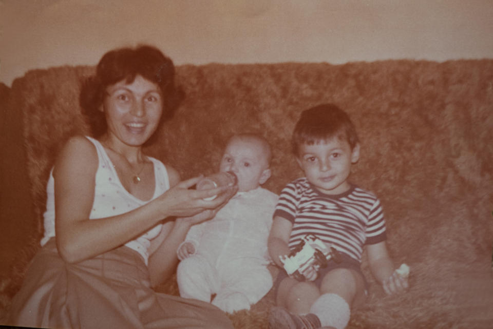 A woman sits on a couch holding a baby and a bottle, with a young boy holding a toy seated next to her. Names: unknown