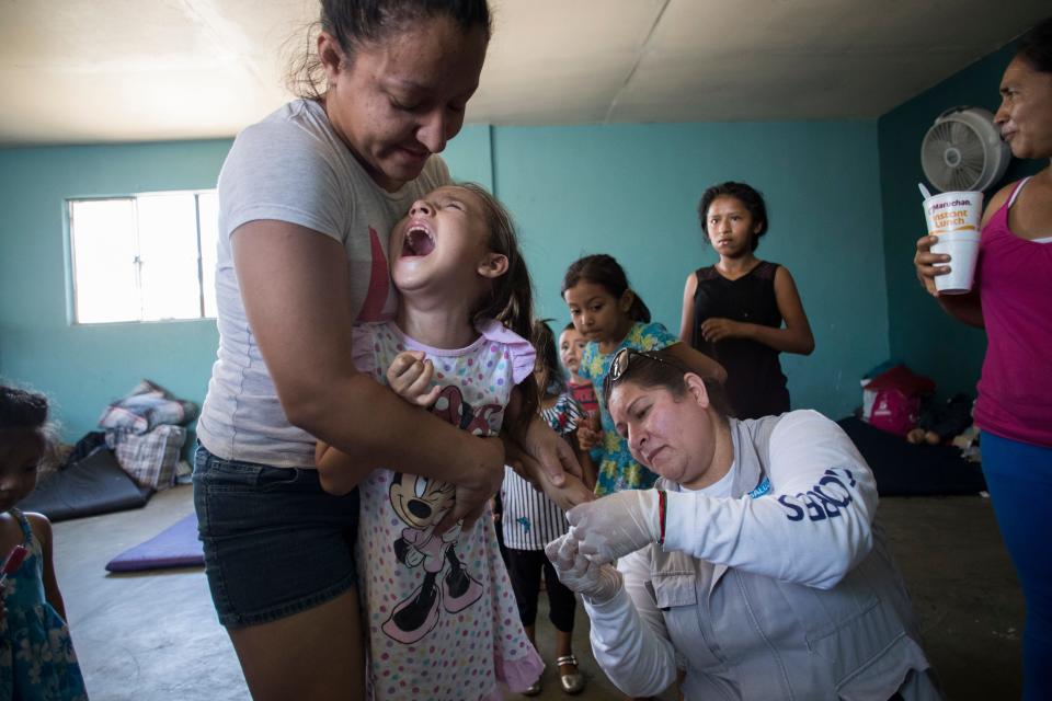 MEXICALI, México –Marisela López, 28, carga en brazos a su hija de 6 años, Maritza Salazar López, mientras Andrea Delgado, representante del Ministerio de Salud de Baja California, le saca sangre por un dedo el 24 de junio del 2019. Delgado estaba haciendo exámenes para descartar malaria en el regio de inmigrantes Casa de Ayuda Alfa y Omega.