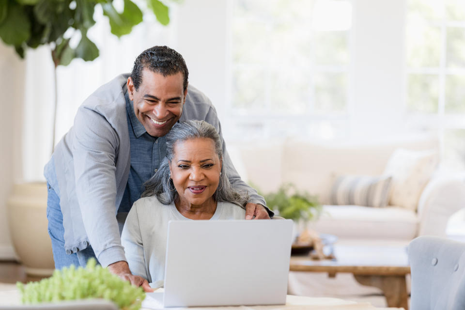 The senior husband stands behind his wife and looks over her shoulder at something displayed on the laptop screen.