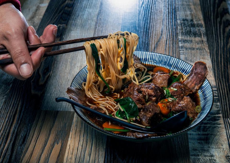 A hand grabs noodles with chopsticks from a bowl