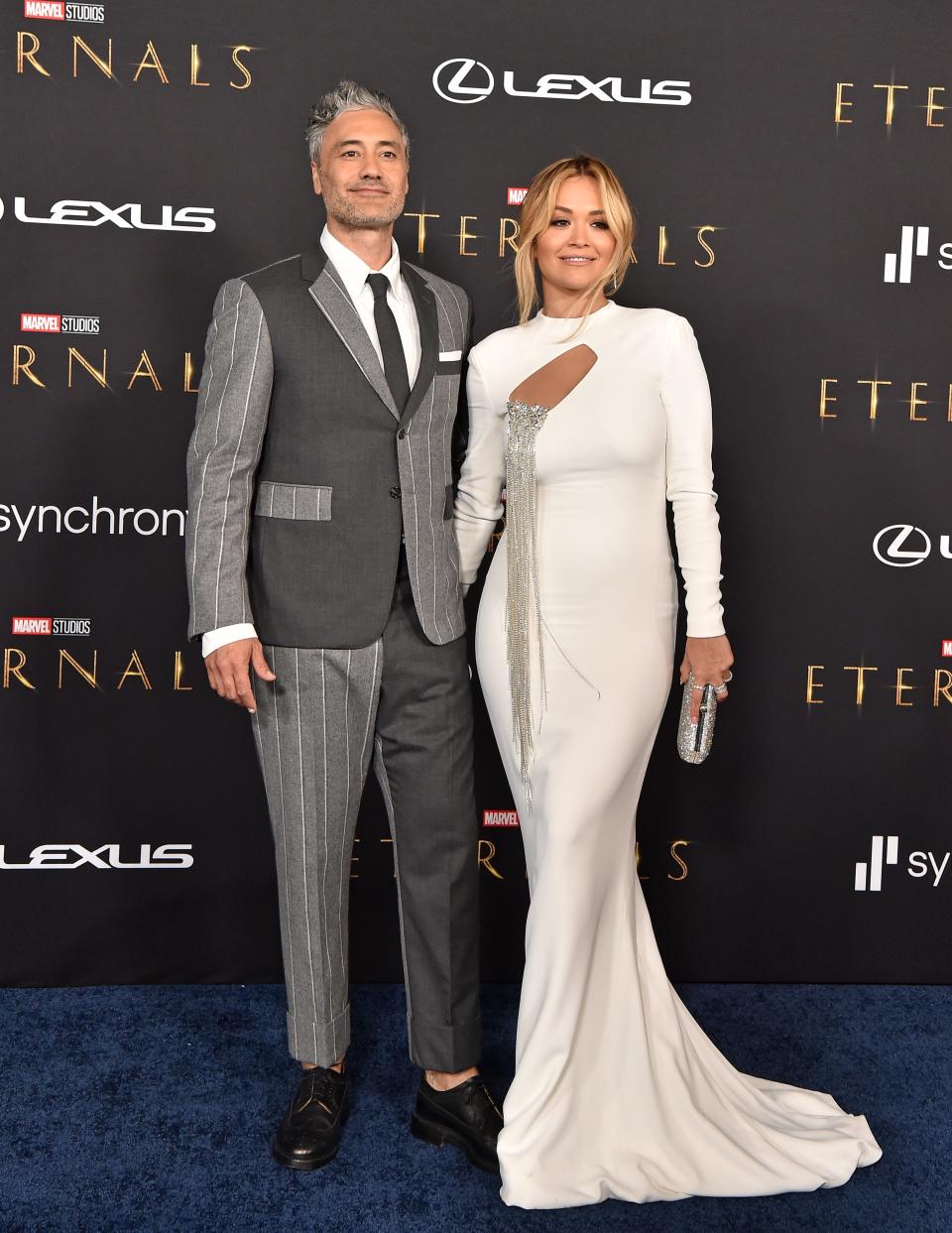 Taika Waititi in a grey suit and Rita Ora in a white dress