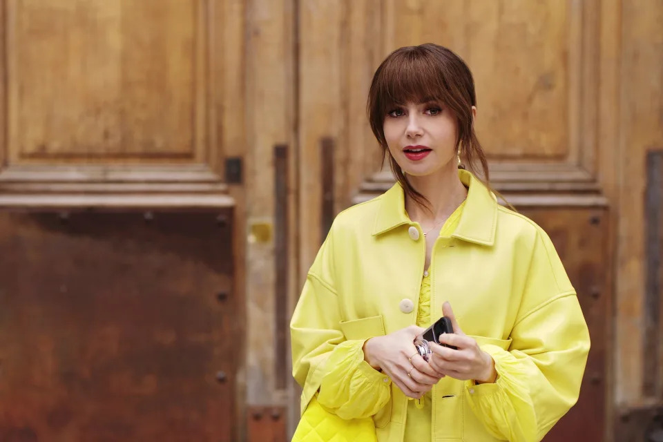 Lily Collins stands in front of a wooden backdrop, wearing a chic, oversized yellow jacket in "Emily in Paris"