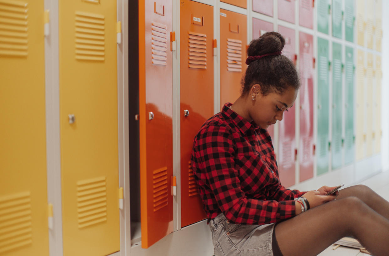 Girl sitting on floor looking at phone held in lap with back against row of multi-colored school lockers.