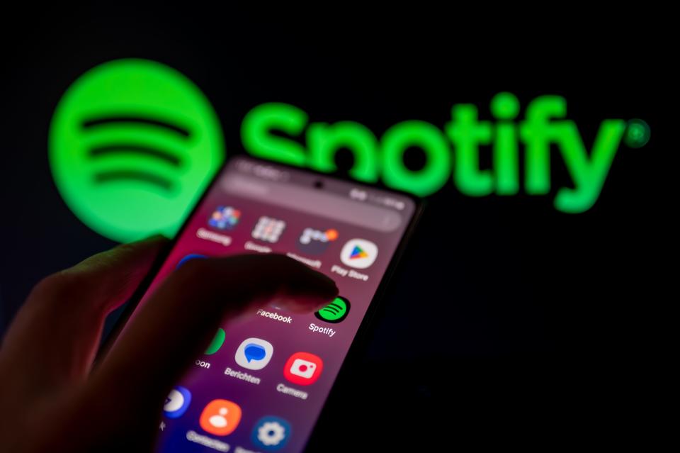 Finger taps the Spotify app on a phone with the Spotify logo blown up in the background.