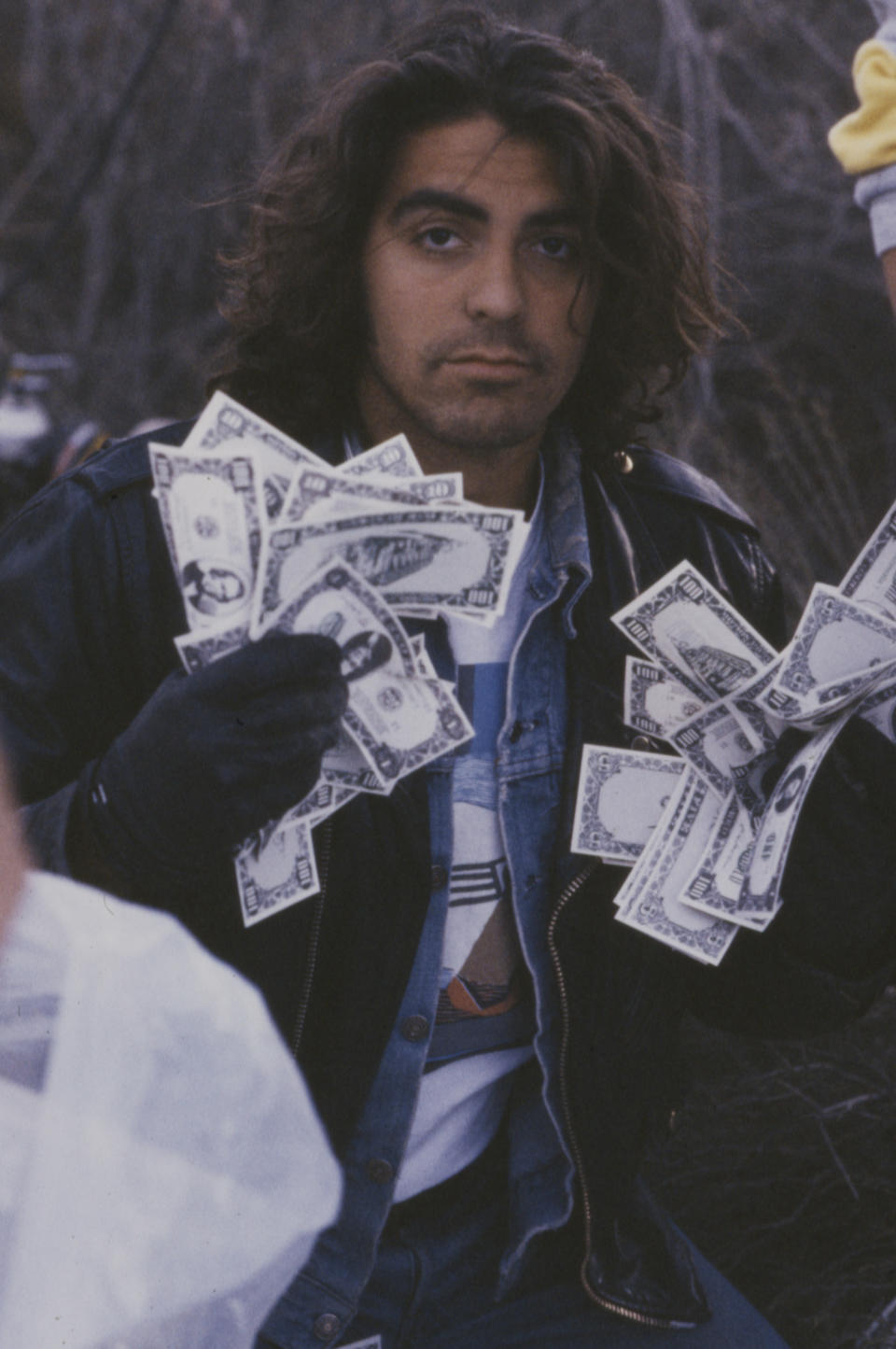 A person wearing a leather jacket over a denim jacket is holding multiple dollar bills in both hands outdoors