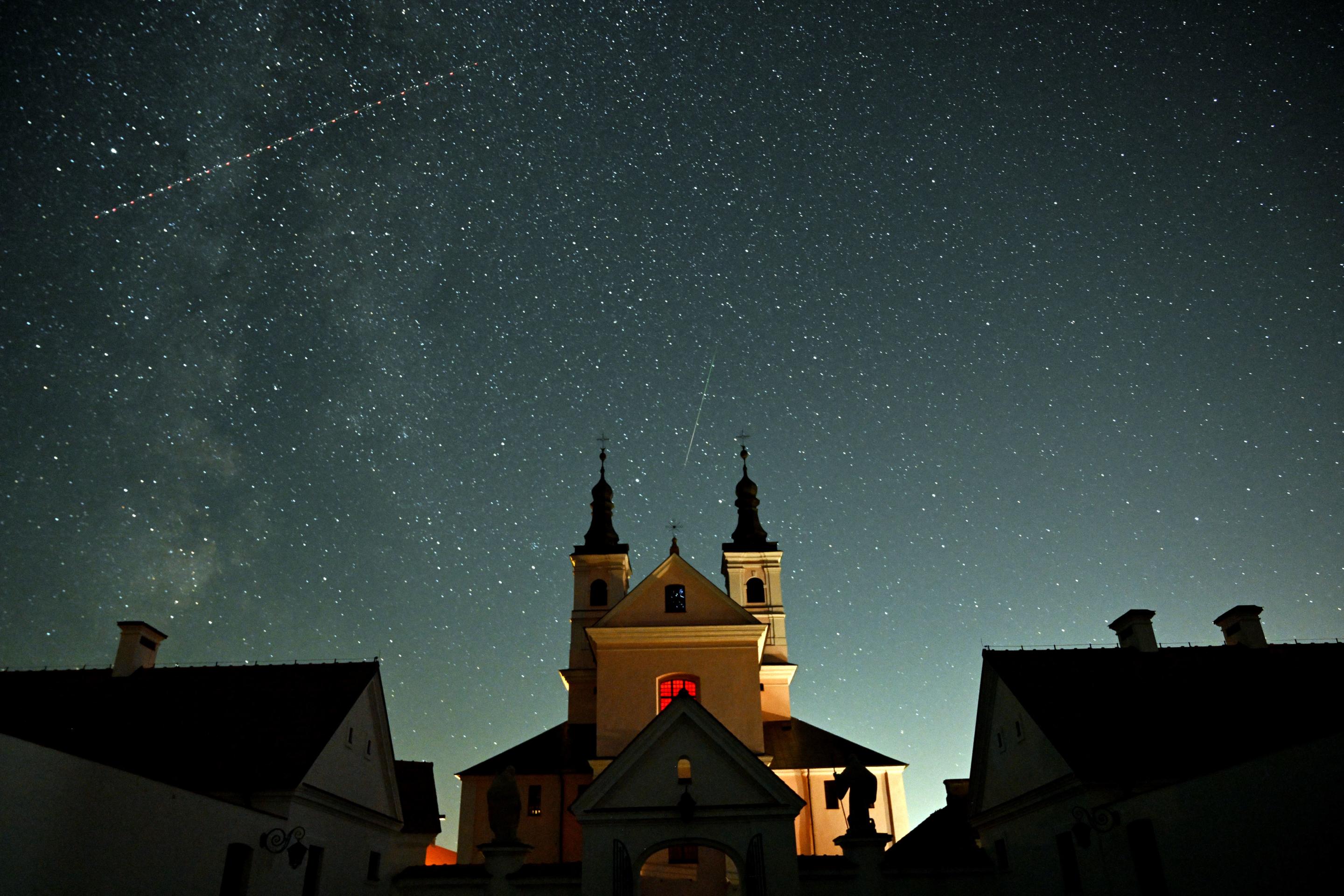 A meteor, a középpont, mint egy műhold, amely átszeli az éjszakai égboltot egy kolostor felett, a lengyelországi Suwalki régióban.