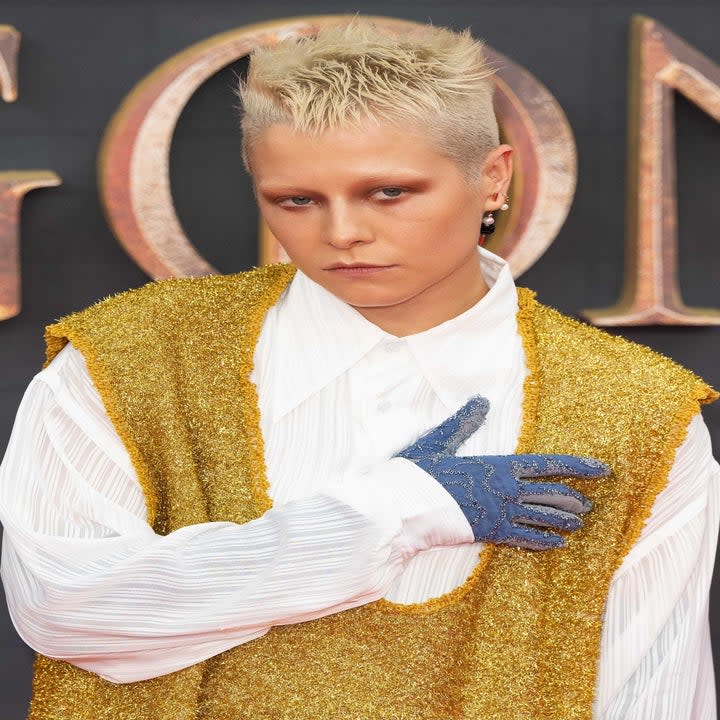 Emma posing on the red carpet for photographers while wearing a long-sleeved dress shirt and a sparkly vest with pants and gloves