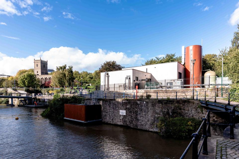 A building housing a heat pump by the water, with a church spire in the background