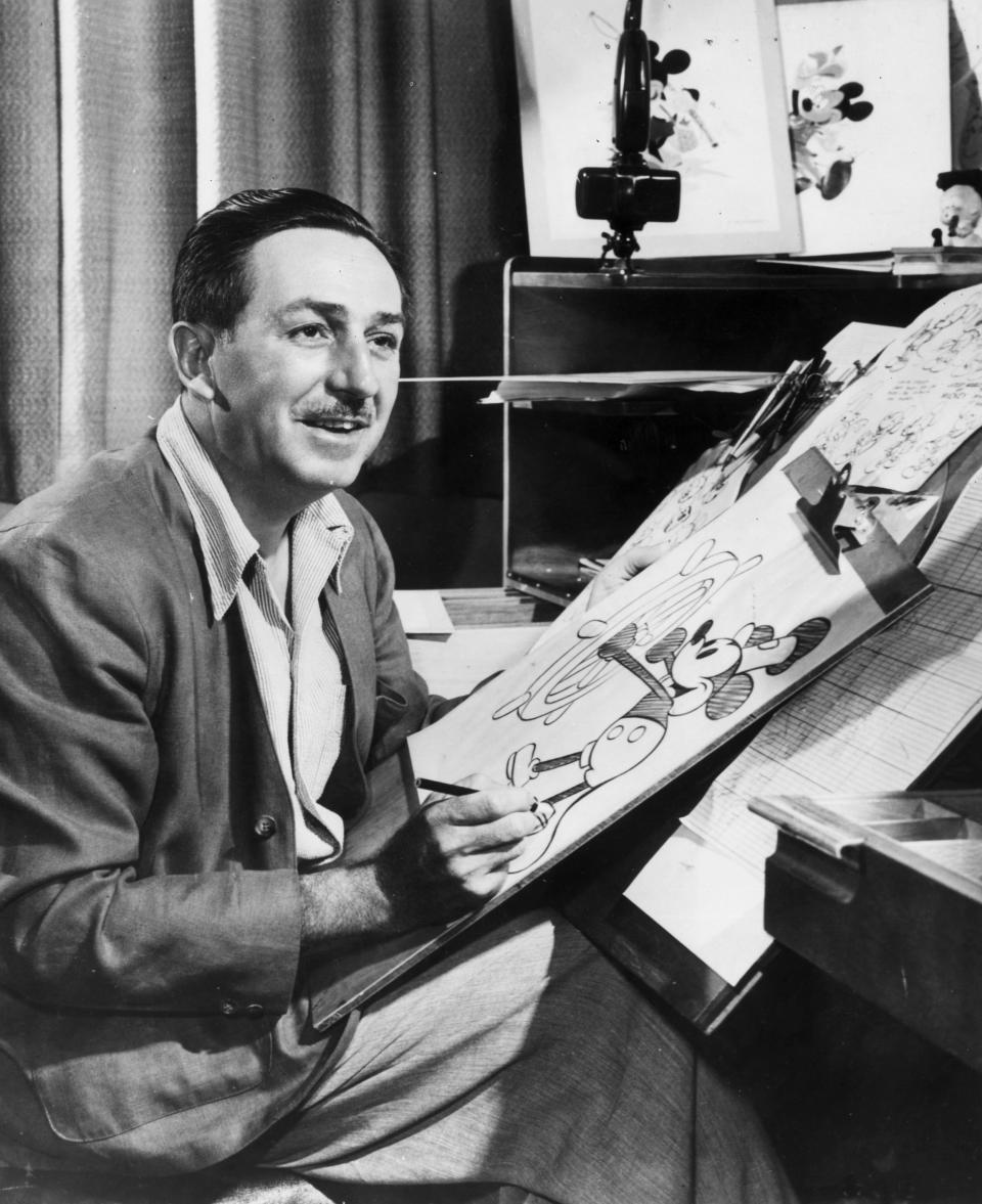 Walt Disney sits at his drawing board in his studio, drawing a sketch of Mickey Mouse.